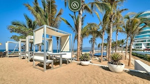 Een strand met witte ligbedden en een overdekte zithoek, omringd door palmbomen en een helderblauwe lucht.