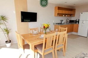 A dining area with a wooden table and chairs, a flat-screen TV mounted on the wall, and a kitchen area with wooden cabinets and appliances. - Ocean View 2BR Condo - Private Terrace - Beach (Nuevo Nayarit)