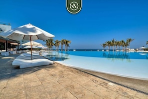 Área de piscina com espreguiçadeiras e guarda-sóis, palmeiras e céu azul límpido.