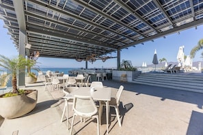 Um pátio externo com mesas e cadeiras, área coberta com painéis solares e vista para o mar e montanhas.