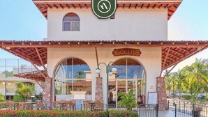 A building with a sign reading 'MACALLANES,' a covered outdoor seating area, and a landscaped entrance. - 2 BR Condo in Hotel - Beach - Pools - Kids Club (Puerto Vallarta)