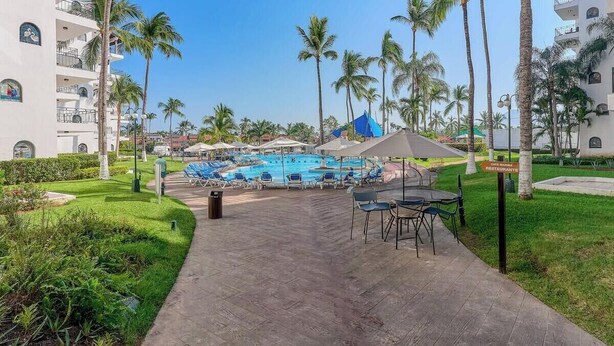 A resort pool area with lounge chairs, palm trees, and a sign indicating a restaurant. - 2 BR Condo in Hotel - Beach - Pools - Kids Club (Puerto Vallarta)