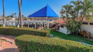 A covered outdoor seating area with a blue roof, surrounded by well-maintained hedges and palm trees. - 2 BR Condo in Hotel - Beach - Pools - Kids Club (Puerto Vallarta)