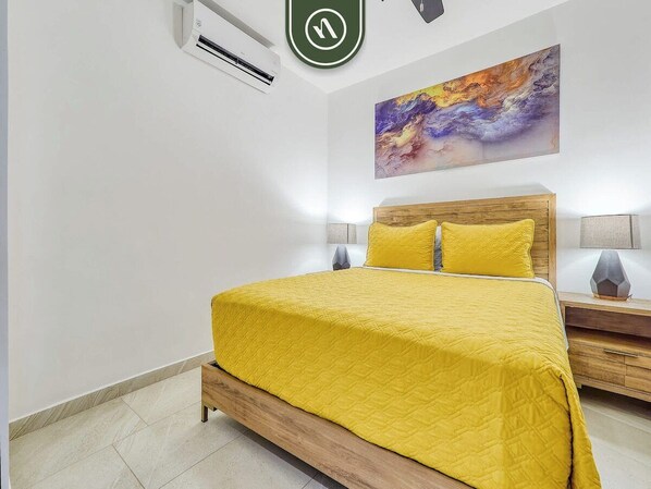 A bedroom with a bed, a nightstand, a wall-mounted air conditioner, and a painting. - Luxury 2BR Condo in Romatic Zone - Rooftop Pool (Puerto Vallarta)