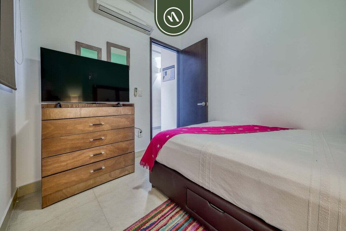 Une chambre avec une commode en bois, un lit recouvert d’une couverture rose et une porte sur laquelle est apposée une pancarte.