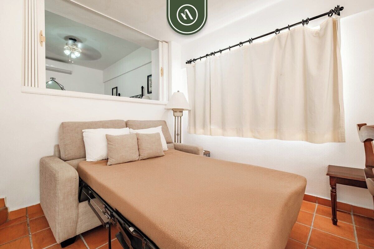 A compact room with a bed, a small table, a chair, a window with curtains, a mirror, and a ceiling light fixture.
