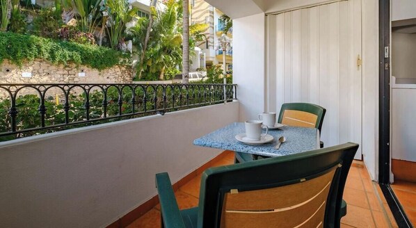 Un balcon avec une table et des chaises, un mur de pierre orné de verdure et une vue sur des palmiers et des immeubles.