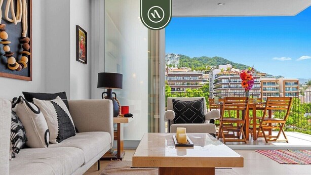 A modern living room with a sofa, coffee table, and a view of a cityscape through large windows. - Beautiful 2 BR Condo Amazing Rooftop Pool - Gym (Puerto Vallarta)