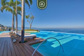 Une piscine à débordement avec une terrasse en bois, des chaises longues et des palmiers.