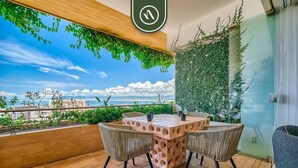 A balcony with a wooden table and chairs, a view of the city, and a green wall. - Luxury 1BR Condo With Private Terrace - Ocean View (Puerto Vallarta)