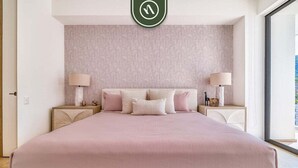 A bedroom with a large bed, two bedside tables with lamps, a patterned wallpaper, and a view through a window. - Luxury 1BR Condo With Private Terrace - Ocean View (Puerto Vallarta)