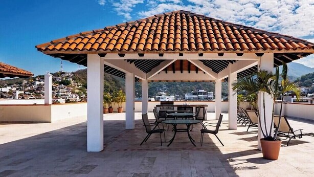 A covered outdoor seating area with a tiled roof, surrounded by potted plants, overlooking a residential area. - Ocean View 2Br Condo-balcony-beach-pool - 206 (Puerto Vallarta)