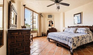 A bedroom with a bed, a wooden dresser, a ceiling fan, a window with curtains, and a wall-mounted air conditioning unit.