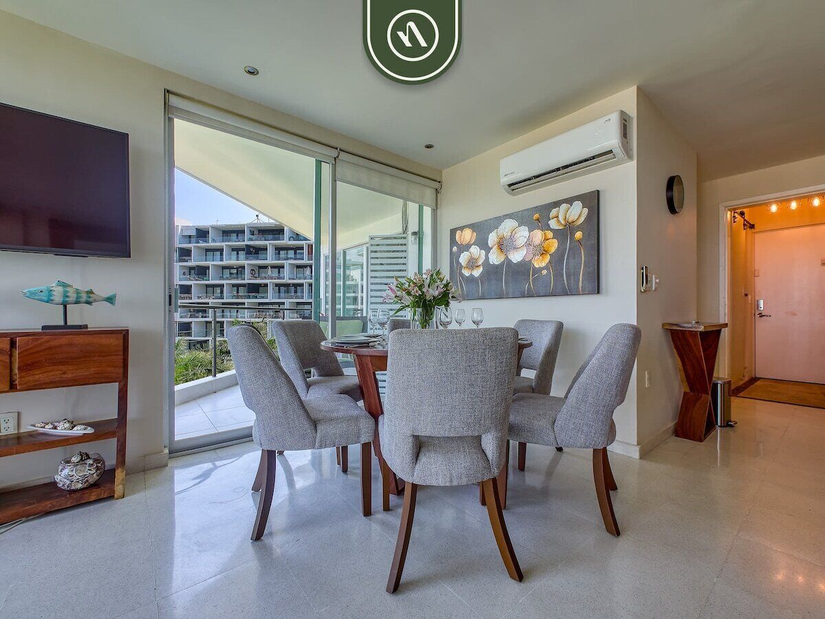 A modern dining area with a round table and grey upholstered chairs, a flat-screen TV, and a large window offering a view of buildings.