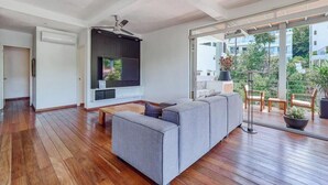 Un salon moderne avec un canapé gris, un plancher en bois, un ventilateur de plafond et une grande fenêtre donnant sur un paysage verdoyant.