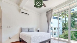 Un dormitori amb llit, ventilador de sostre, unitat de climatització, finestra amb cortines i vista a edificis i vegetació exterior.