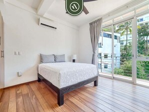 Een slaapkamer met een bed, een plafondventilator, een airconditioningapparaat, een raam met gordijnen en uitzicht op gebouwen en groen.