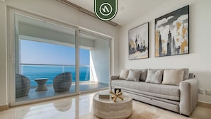 A modern living room with a grey sofa, two side tables, and a round coffee table. There are two framed artworks on the wall and a sliding glass door leading to a balcony with a view of the sea. - Oceanview 1 BR Condo With Pools and Beach Access (Puerto Vallarta)