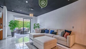 A modern living room with a sectional sofa, a matching ottoman, and a large window offering a view of a green outdoor area. - Modern 2BR Condo in Old Town - Private Terrace (Puerto Vallarta)