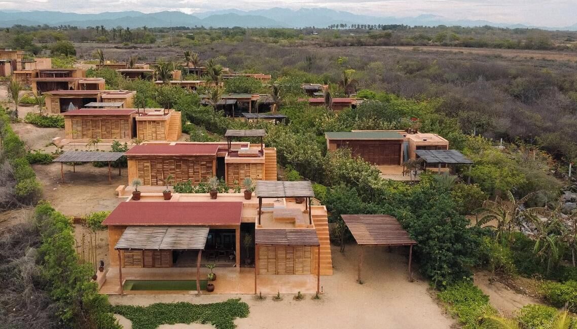 A cluster of wooden cabins with flat roofs, surrounded by greenery and mountains in the background.