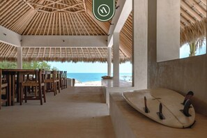 En avslappnad lounge vid stranden med ett halmtak, trästolar och ett ställ för surfbrädor.