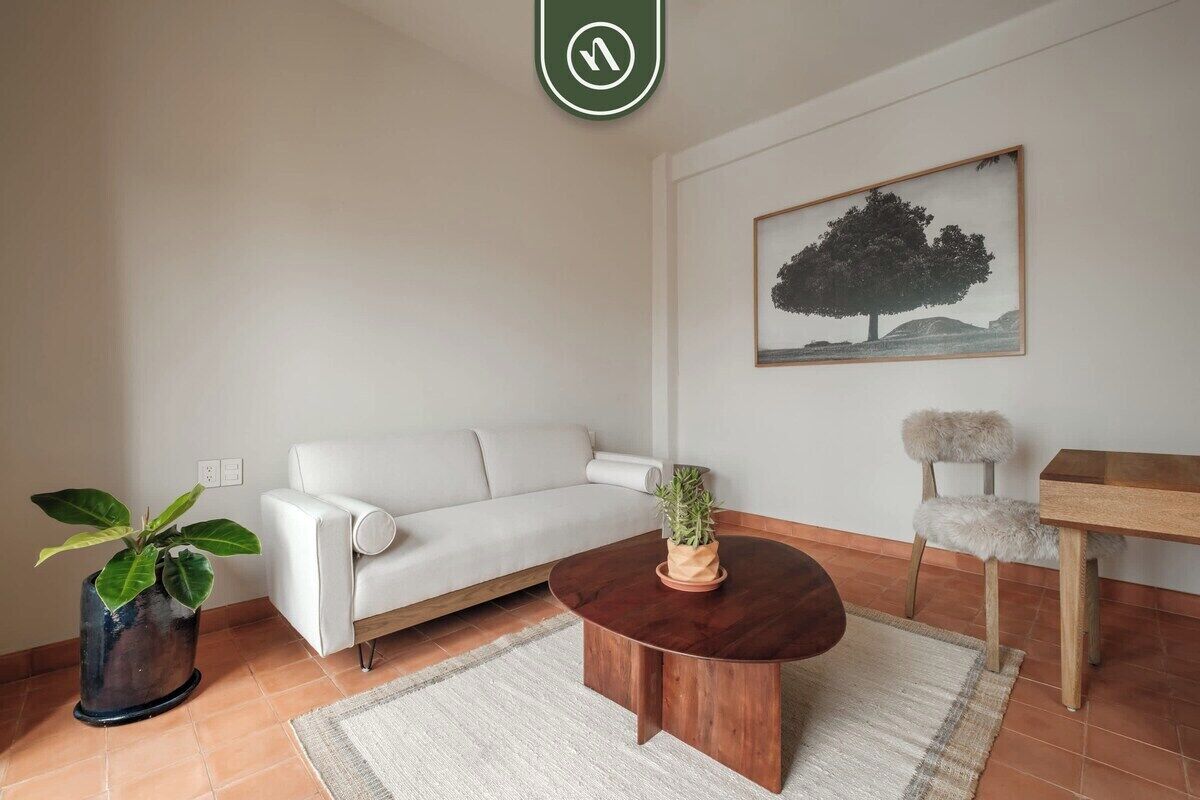 A living room with a white sofa, a round wooden coffee table, a potted plant, a chair, and a painting on the wall.