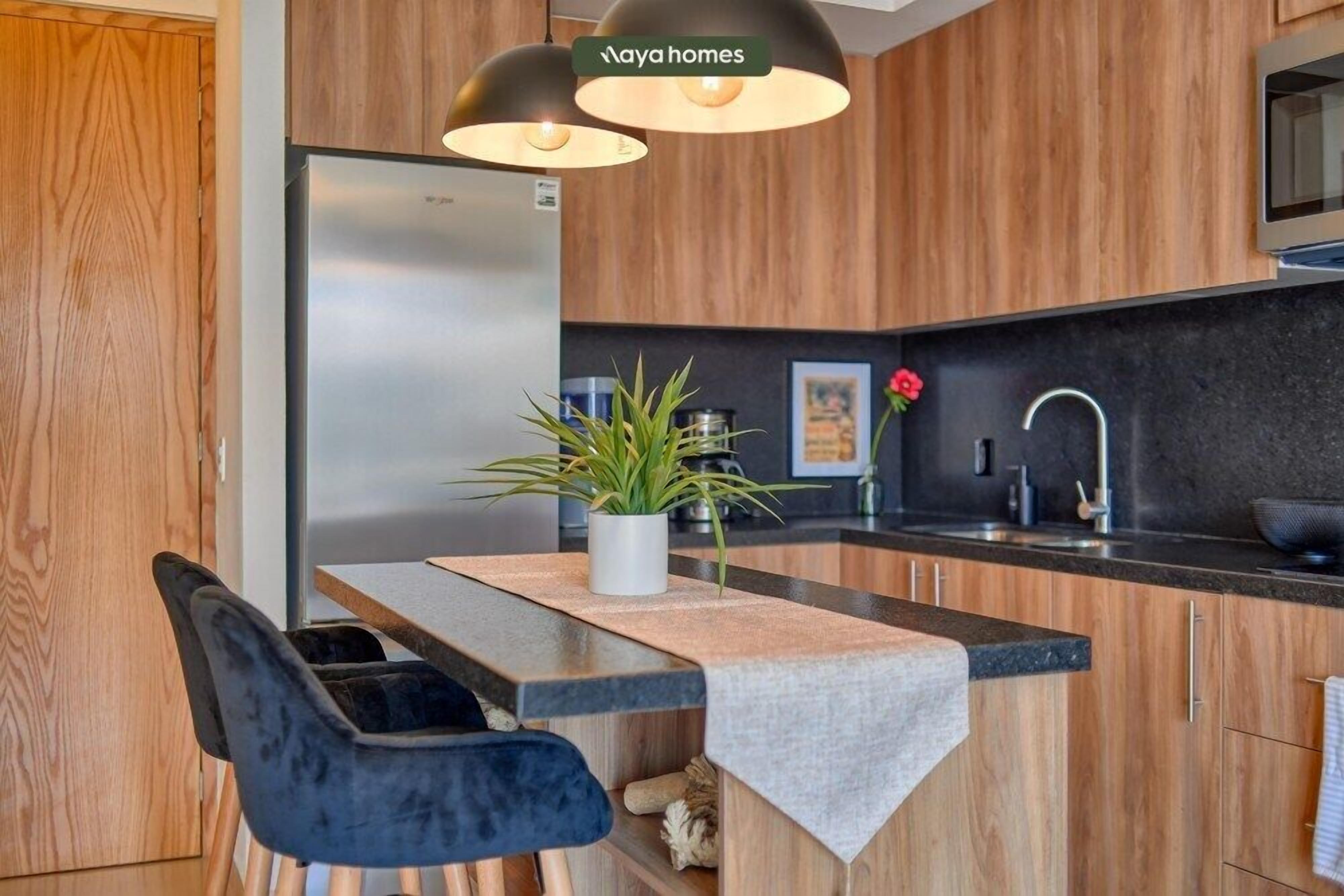Une cuisine moderne avec des meubles en bois, un réfrigérateur en acier inoxydable, un plan de travail noir et une plante en pot.