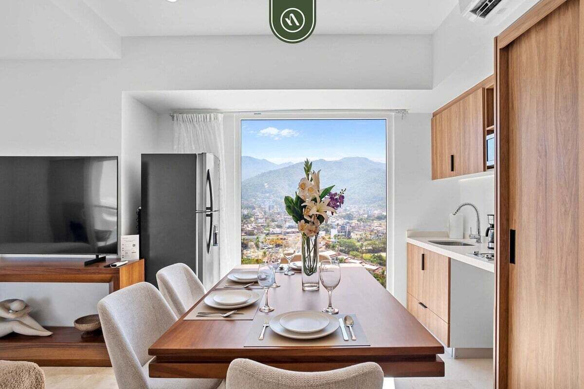 A modern dining area with a wooden table set for four, a vase of flowers, and a city view through large windows.