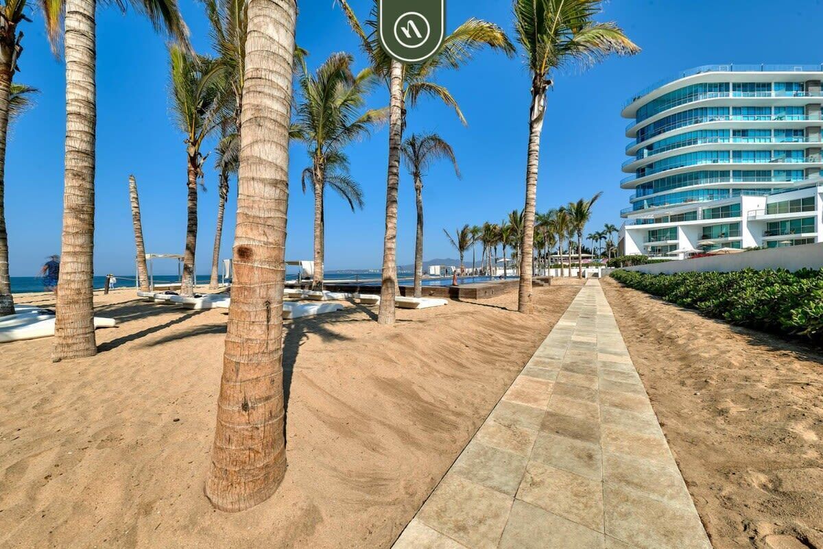 Ett hotell vid stranden med palmer, en sandpromenad och en modern byggnad med balkonger.