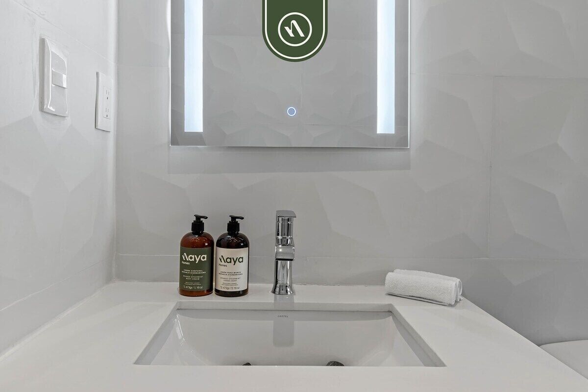 A bathroom with a white sink, two bottles of Ayya body wash, a mirror with a green logo, and a white towel on the counter.