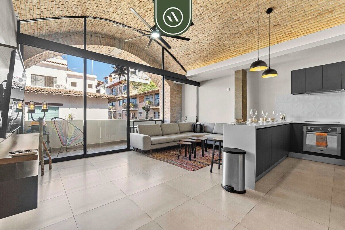 A modern kitchen with a built-in oven, a dining area with a sofa and chairs, and a view of the city through large windows.