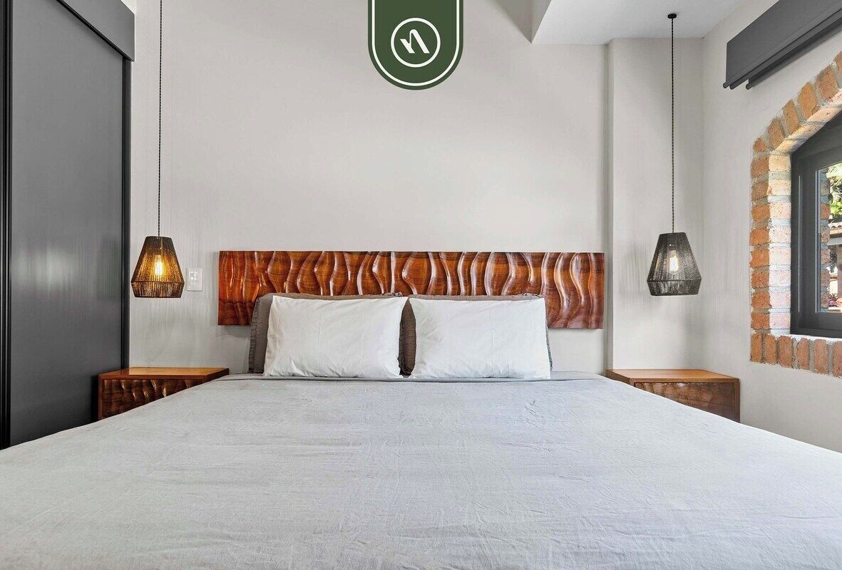 A modern hotel room with a large bed, wooden headboard, two bedside tables, and hanging lamps.