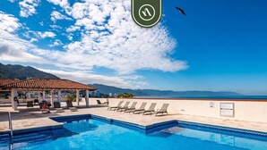 A pool with lounge chairs and a covered area, overlooking a beach and mountains.