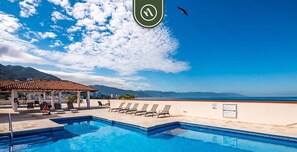 A pool with lounge chairs and a covered area, overlooking a beach and mountains.