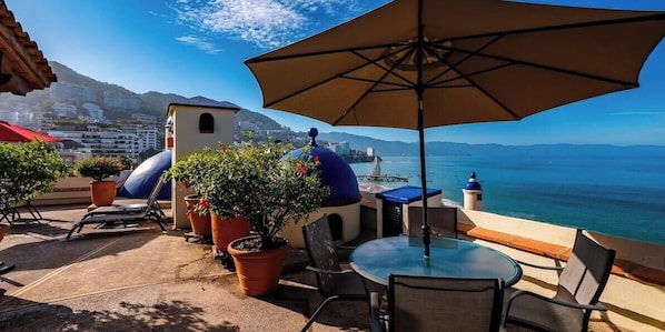 Eine Terrasse im Freien mit Tisch und Stühlen, Kübelpflanzen und Blick auf das Meer.
