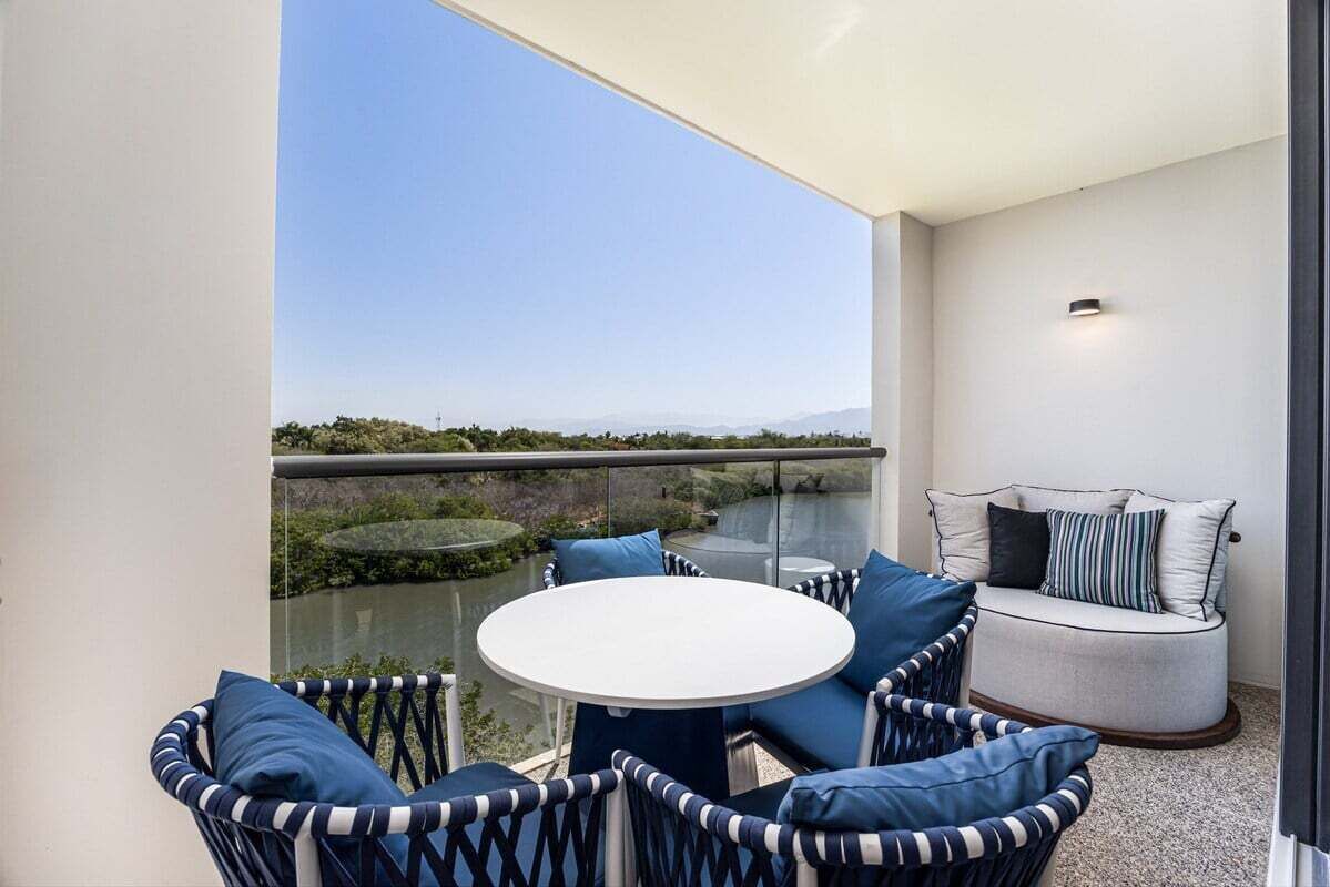 Een balkon met een ronde tafel en rieten stoelen, een gewatteerd bankje en uitzicht op een waterpartij en een verafgelegen landschap.