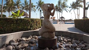 Eine Steinstatue in einem kreisförmigen Wasserspiel, umgeben von Steinen, mit Palmen und blauem Himmel im Hintergrund.