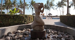 A stone statue in a circular water feature surrounded by rocks, with palm trees and a clear sky in the background. - Mexican Styled 2 BR Condo - Beachfront - Pool (Nuevo Nayarit)