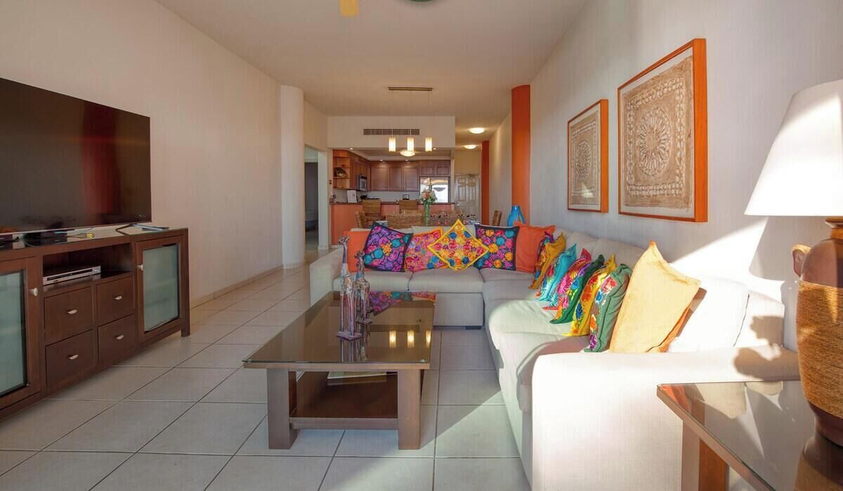 A living room with a sofa, a coffee table, a TV, and a kitchen in the background.