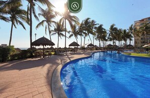 Une piscine bordée de palmiers et agrémentée de parasols, le tout entouré d’une allée pavée en briques.