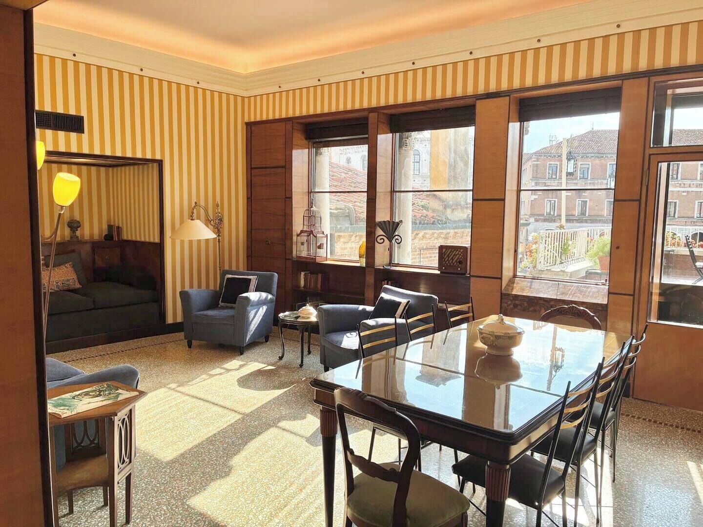 Quarto com mesa de jantar, cadeiras, sofá e janela com vista para edifícios.