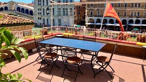 An outdoor terrace with a table and chairs, overlooking a historic building with arches and a tiled roof.