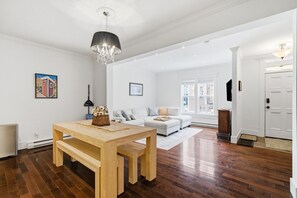 A spacious living room with a wooden dining table, a white sofa, and a chandelier. - Spacious Townhouse | 3 BR, Steps to Water St. (St. John`s)