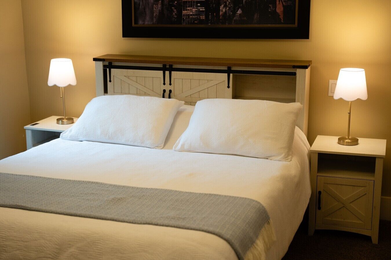 A neatly made bed with two pillows, a bedside table with a lamp, and a nightstand with a lamp.