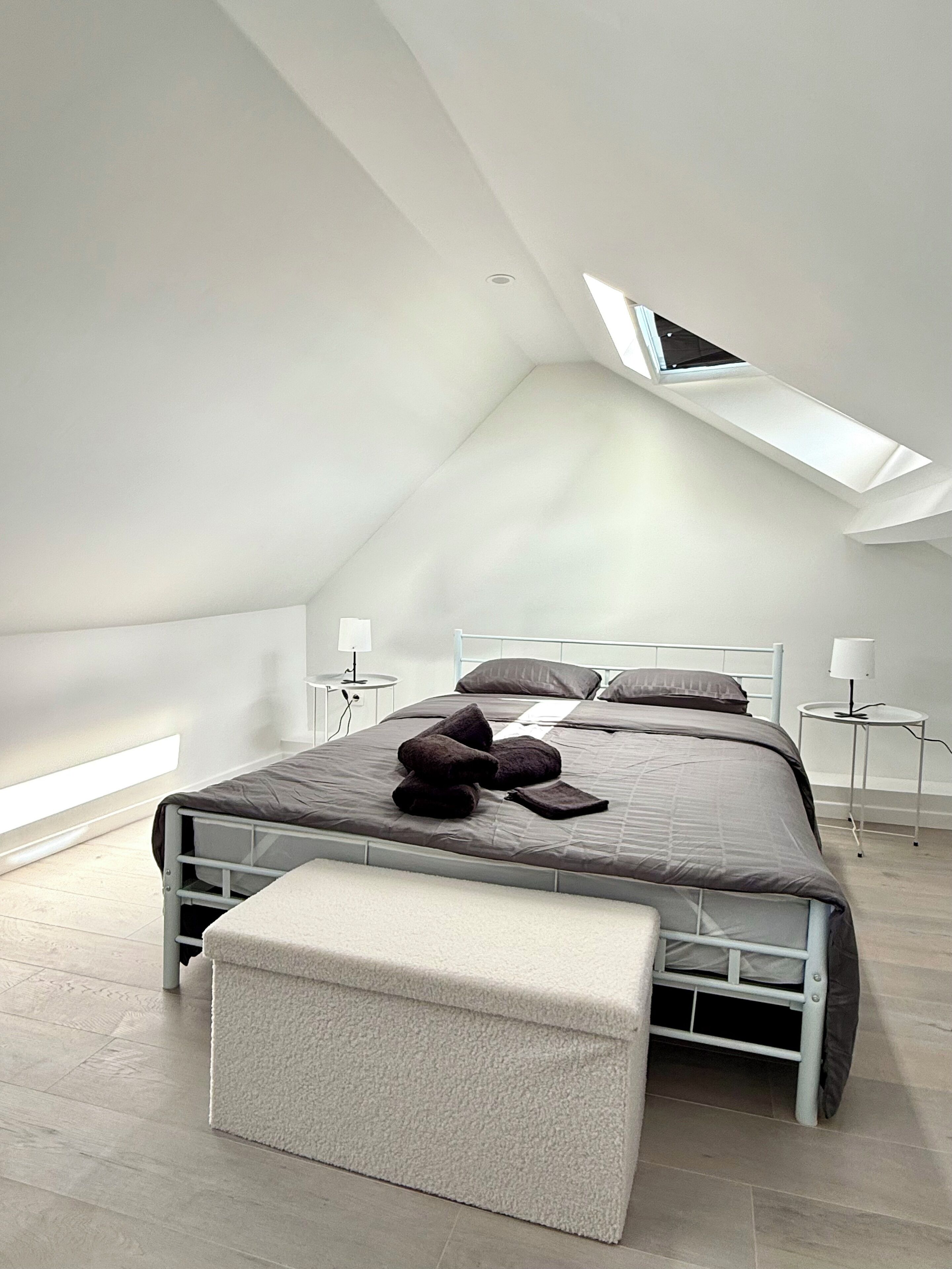 Een moderne slaapkamer met een schuine kap, een bed met een wit frame, een bankje bij het voeteneind en twee nachtkastjes met lampen.