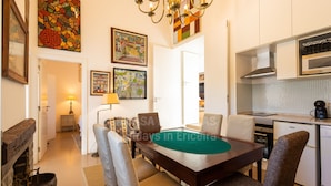 A dining area with a wooden table, chairs, and a green felt tablecloth. There are framed artworks on the walls, a chandelier, and a kitchen area with modern appliances.