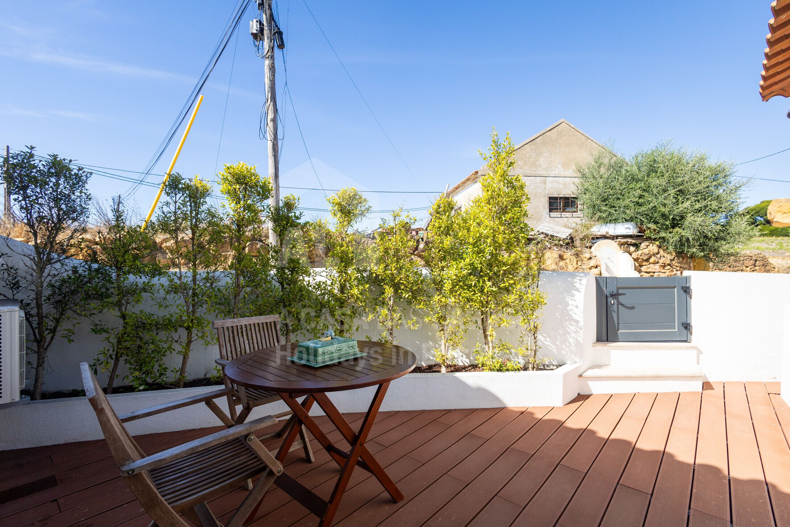 A wooden deck with a table and chairs, surrounded by greenery and a house in the background.