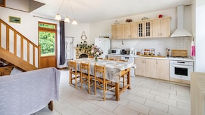 Una cocina con armarios de madera, una mesa de comedor con sillas, una escalera y vistas al exterior con vegetación.