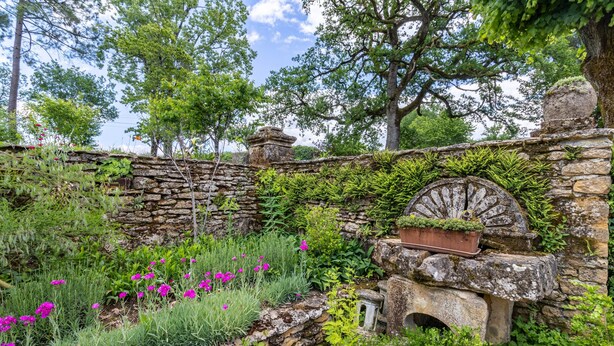 Un muro de piedra con una estructura circular, abundante vegetación y plantas con flores.