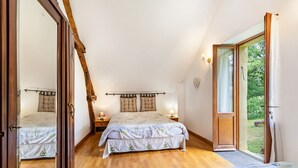 Un dormitorio con una cama grande, otra más pequeña, techo de madera, una mesita de noche, una lámpara y un ventanal corredero a un jardín.